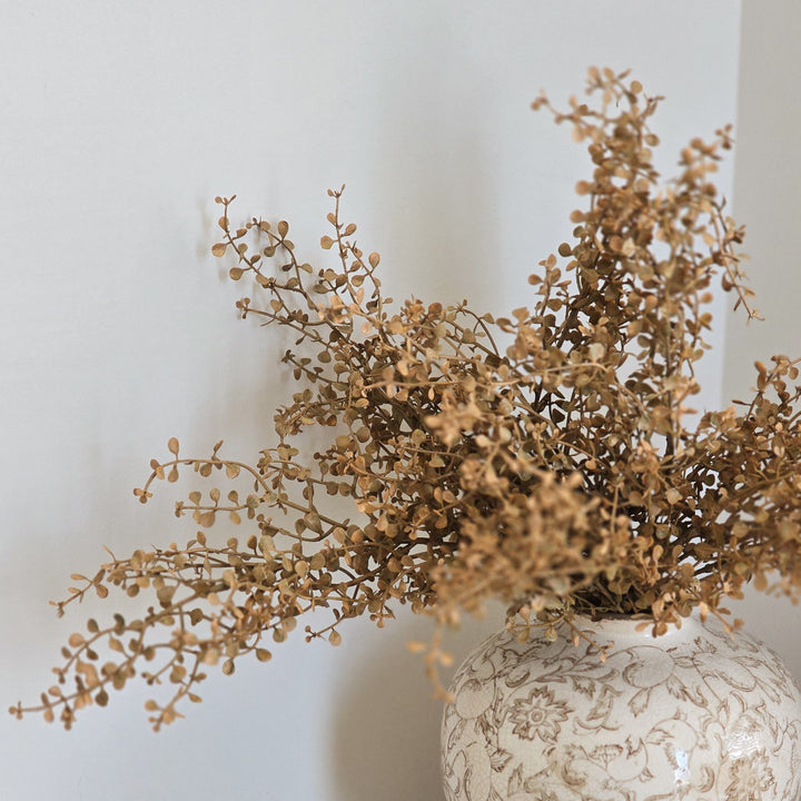 A ceramic vase with a beige and brown leaf pattern, filled with dried brown branches, sitting on a rustic wooden console table next to a small wooden lotus-shaped dish.