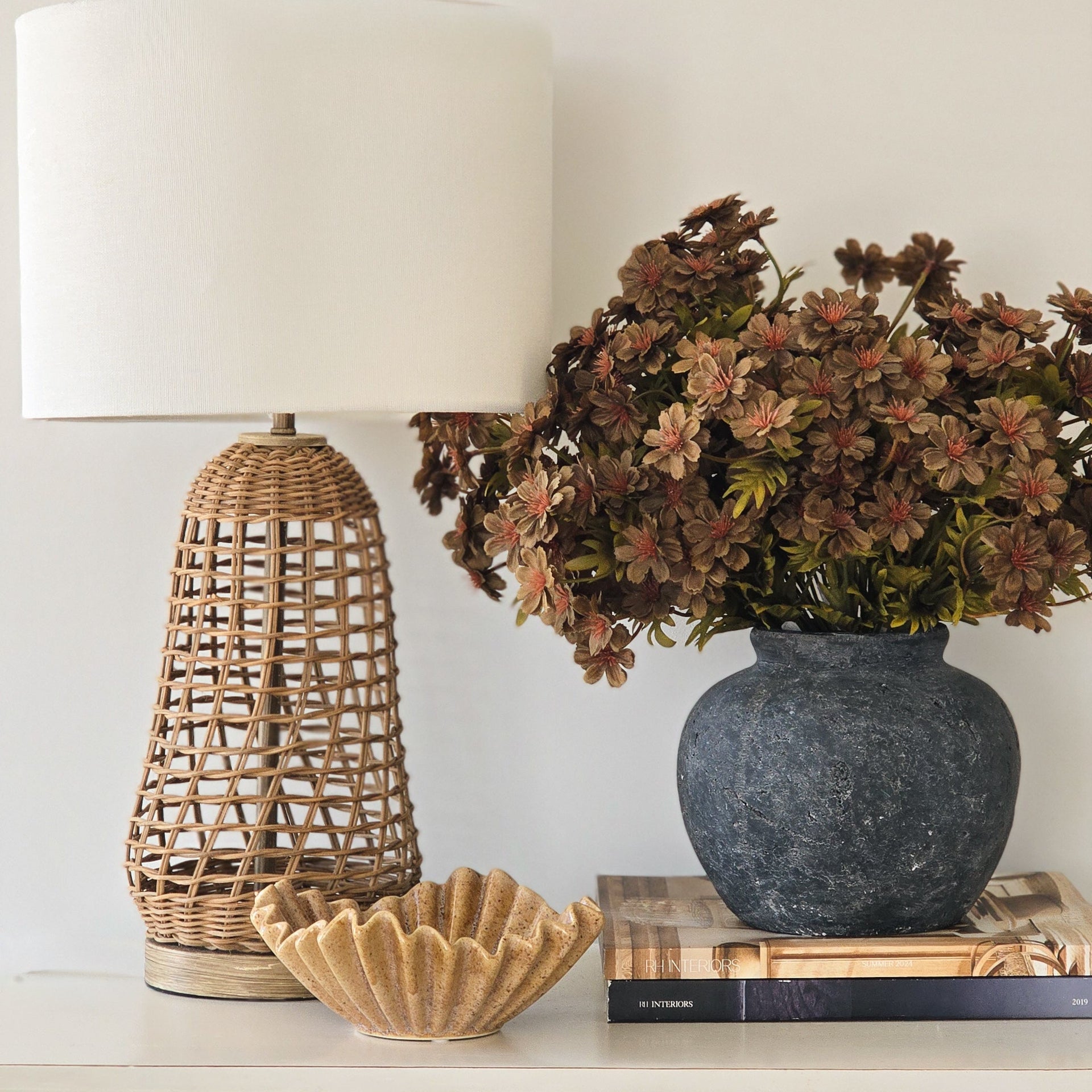 A styled interior scene showing a woven rattan table lamp with a white drum shade, next to a dark blue-gray ceramic vase filled with dried brown flowers, and a small scalloped beige bowl on a white cabinet.