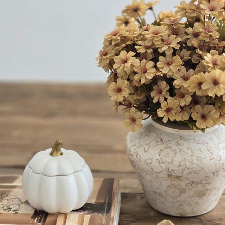 A styled tabletop scene with a ceramic vase filled with tan dried flowers, a small white pumpkin-shaped container on stacked books, and a beige flower-shaped dish on a rustic wooden surface against a light gray background.