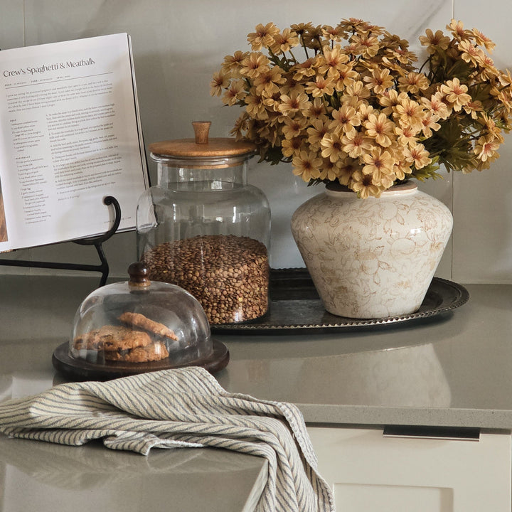 A styled kitchen countertop scene with a ceramic vase holding dried beige flowers, a glass jar filled with brown grains, a small glass-domed pastry cover, and a striped cloth napkin on a gray stone counter.