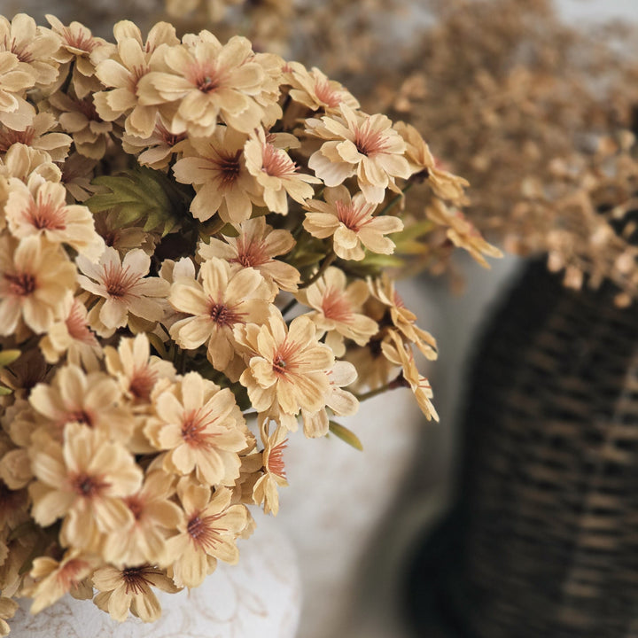 A close-up of dried beige flowers with soft pink centers arranged in a textured white ceramic vase on a round tray, with a darker woven vase blurred in the background.