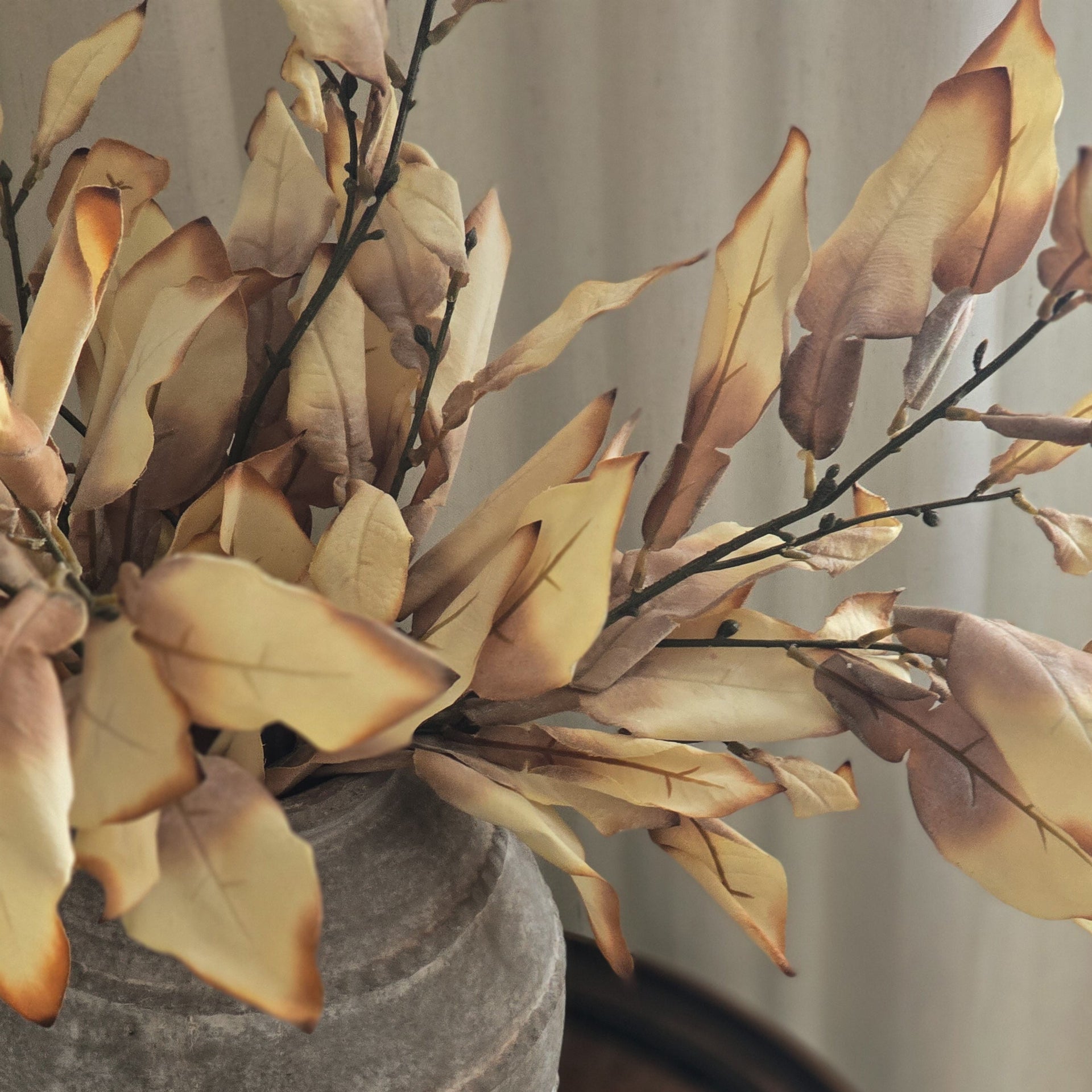 A small white candle in a clear glass holder sits on a dark wooden side table next to a gray ceramic vase filled with dried beige and brown leaves, against a soft neutral curtain background.