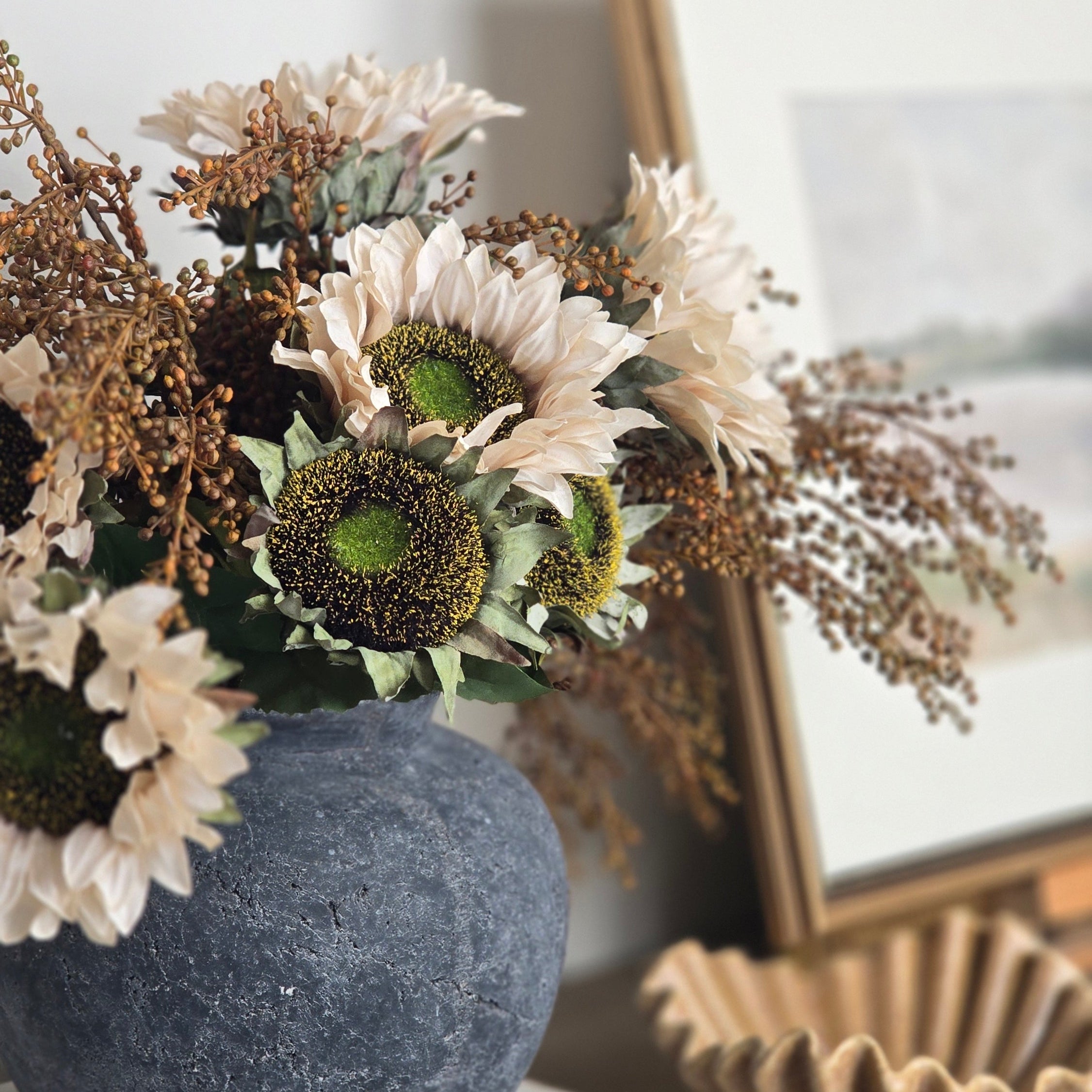 A dried flower arrangement in a textured dark blue-gray ceramic vase, styled on a wooden console table next to a framed picture and a decorative shell bowl.