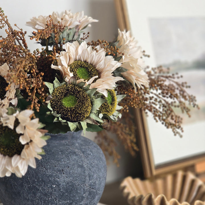 A dried flower arrangement in a textured dark blue-gray ceramic vase, styled on a wooden console table next to a framed picture and a decorative shell bowl.