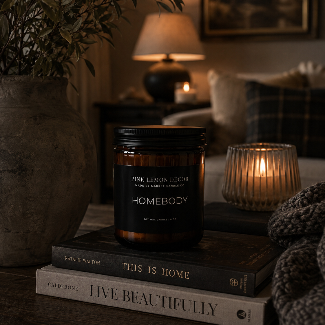 A Homebody scented candle in an amber glass jar with a black lid sits on stacked books on a wooden table in a cozy, dimly lit living room setting with a lit candle holder and warm ambient lighting.