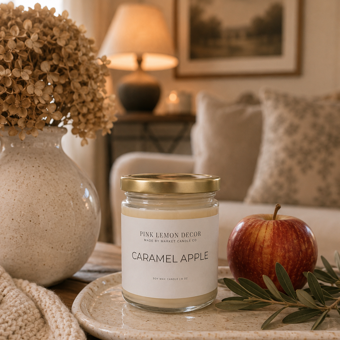 A glass jar candle with a gold lid and white label reading 'PINK LEMON DECOR' and 'CARAMEL APPLE' sits on a tray in a cozy living room setting, styled with a red apple, greenery, and a ceramic vase of dried flowers.