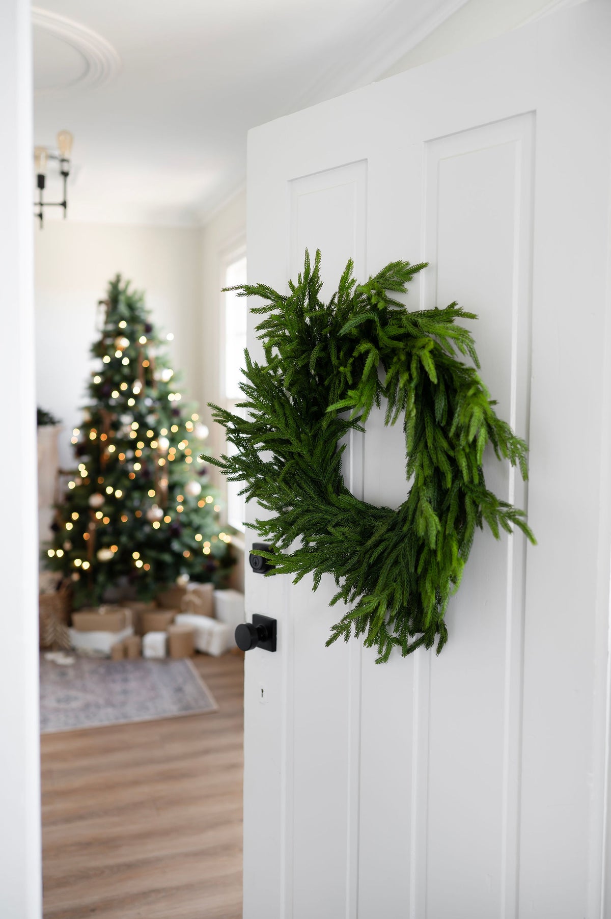 A lush green artificial pine wreath hangs on a white paneled door, with a softly blurred decorated Christmas tree and holiday home interior visible in the background.