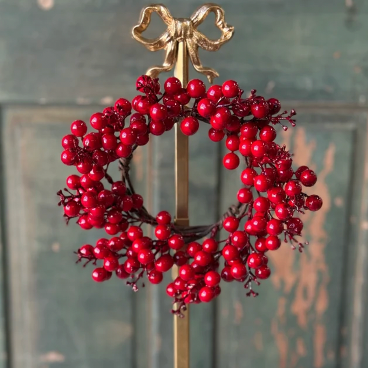 A decorative holiday wreath made of glossy red berries on a gold metal stand, set against a distressed green wooden door background.