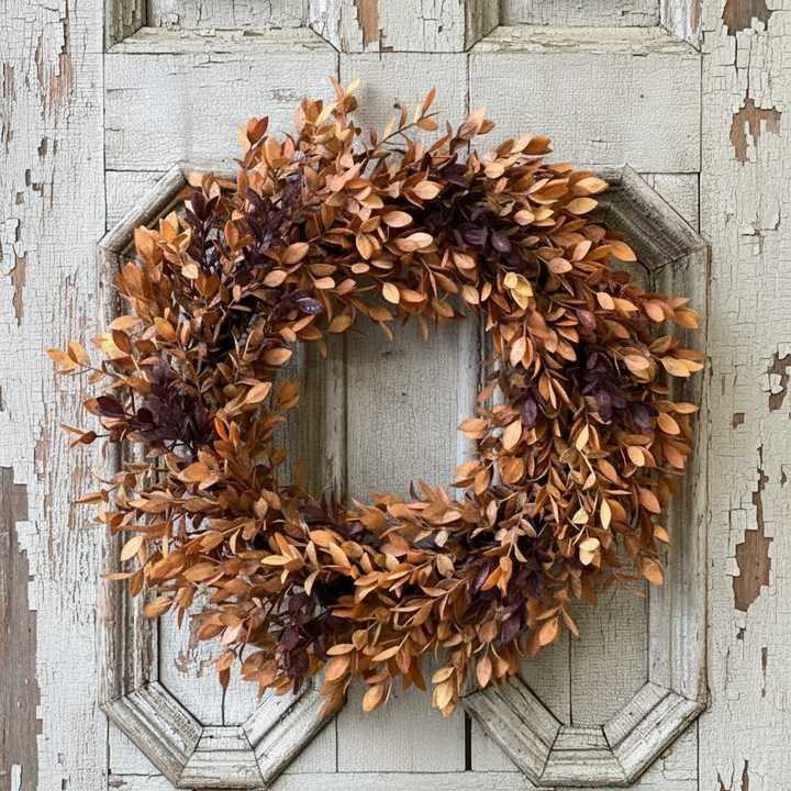 An autumn-themed artificial leaf wreath with warm orange, brown, and deep burgundy leaves, displayed on a distressed white wooden door.