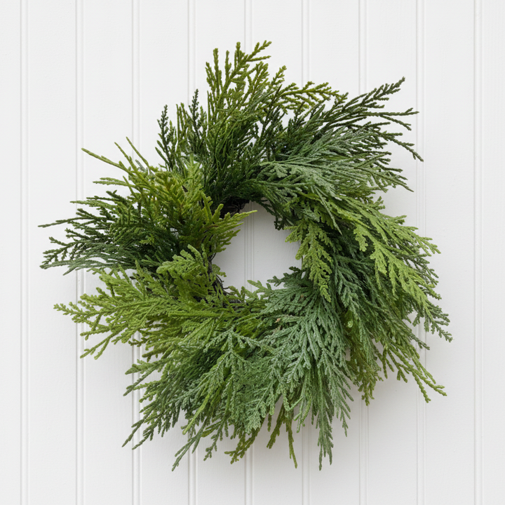 A green artificial cedar wreath with dense, layered needle-like foliage, isolated on a solid white background.