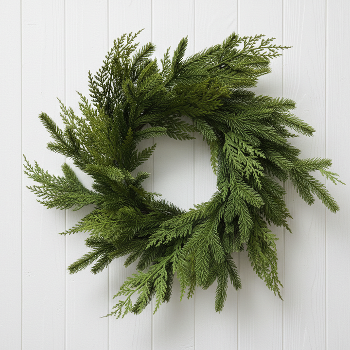 A green artificial pine or cedar wreath with layered, feathery foliage, displayed against a white-painted brick wall.