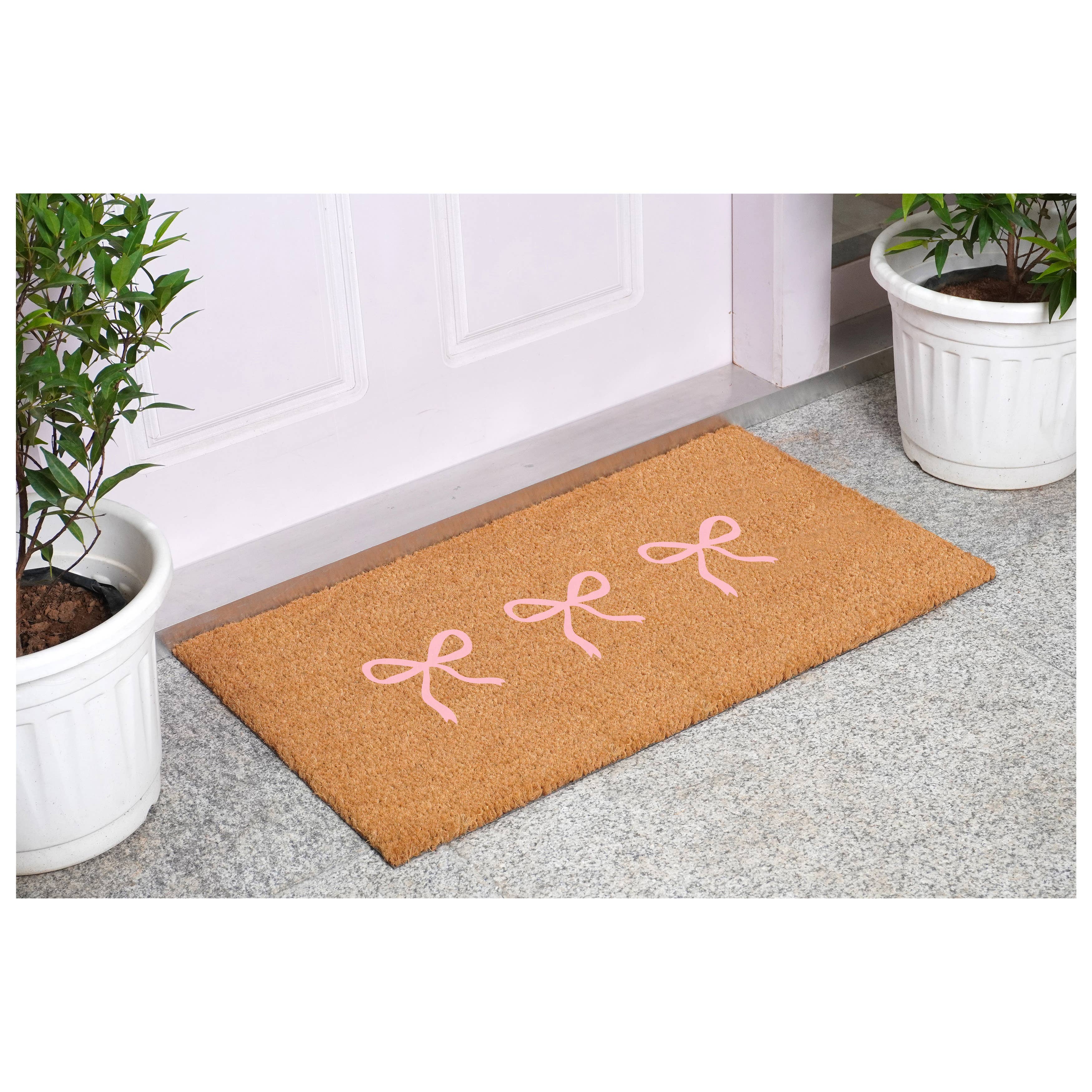 Doormat with pink bows on a gray floor next to a white door.